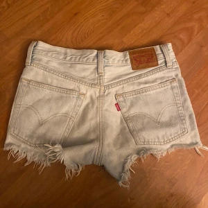 Levi's jeansshorts - Snygga ljusblå jeansshorts från Levi's 501 med medelhög midja och råa fransade kanter. Klassisk femficksmodell med knappgylf och ikonisk läderpatch bak. Perfekta för sommaren och ger en avslappnad vibe till din outfit. Sänker pris vid snabb affär
