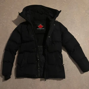 Svart pufferjacka från Zavetti Canada - Snygg svart pufferjacka från Zavetti Canada med huva och fuskpäls. Jackan har dragkedja, stora fickor och patch på ärmen. Pälsen ingår och har varit använd i knappt två år och i makalöst skick.