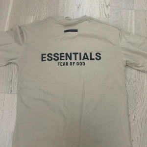 Beige Essentials Fear of God t-shirt - Snygg beige t-shirt från Essentials Fear of God med svart logga tryckt på ryggen. Klassisk rund halsringning och korta ärmar. Tillverkad i mjuk bomull för en skön och avslappnad vibe. Perfekt för dig som gillar streetwear och minimalistisk stil. Produkten har aldrig använts och är helt nytt. Pris kan diskuteras.