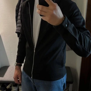 Svart jacka med dragkedja från Jack & Jones -  Svart jacka från Jack & Jones med hel dragkedja framtill och klassisk krage. Jackan har två bröstfickor och långa ärmar med knapp vid ärmslut. Perfekt för en clean och enkel look.