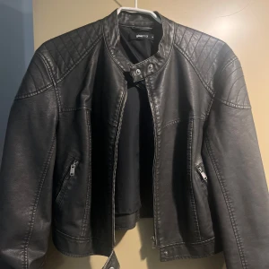 Mörkgrön skinnjacka från Gina Tricot - Snygg mörkgrön skinnjacka från Gina Tricot med quiltade detaljer på axlarna och dragkedjor vid fickorna. Jackan har en croppad passform, ståkrage med tryckknappar, raka ärmar och är helt oanvänd.