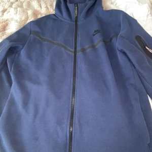 Blå Nike tech fleece - Snygg blå zipjacka från Nike med hel dragkedja och svart Nike-logga på bröstet. Jackan har en sportig look med svarta detaljer över bröstet och en ficka med dragkedja på ärmen. Perfekt för dig som gillar streetwear och sportig stil.