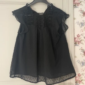 Svart blus med spets från  - Svart blus med transparenta volangärmar och broderad spets upptill. Prickig struktur i tyget och luftig passform. Inte lika bred som den ser ut! Sitter bra men inte i bästa skick!