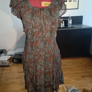 Blommig kortklänning - Supersöt kortklänning med blommigt mönster i brunt, blått och beige. Klänningen har korta ärmar, knytning i midjan och vid halsen samt volangdetaljer. Tunt, luftigt material som passar perfekt till varmare dagar.