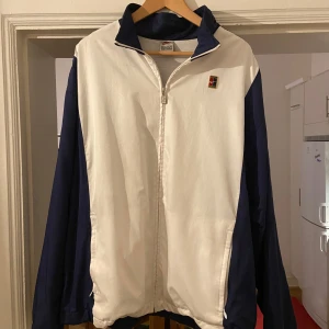 Nike vintage vindjacka XL blå/vit - Retro vindjacka från Nike i storlek XL. Jackan är vit framtill med marinblå ärmar och krage, och har en broderad Nike-logga på bröstet. Klassisk rak passform, hel dragkedja och två sidofickor. Perfekt för dig som gillar sportig och old school stil.