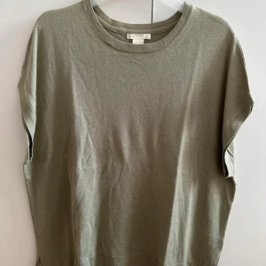 Olivgrön oversized stickad topp med slits - Olivgrön stickad topp med korta ärmar och rund halsringning. Modellen är loose fit och har små slitsar i sidorna nertill för en avslappnad look. Perfekt att styla med jeans eller kjol för en chill vibe