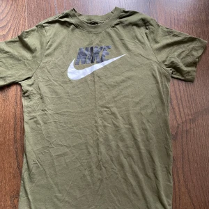 Olivgrön Nike t-shirt med logga - Snygg olivgrön t-shirt från Nike med klassisk stor vit logga på bröstet. T-shirten är i mjuk bomull och har en avslappnad passform med korta ärmar och rund hals. Perfekt för dig som gillar sportig och enkel stil.