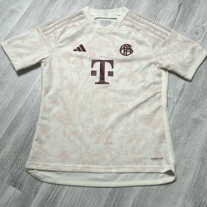 FC Bayern München Adidas fotbollströja - Nästan aldrig använd köpt runt 900kr säljer 350kr.Snygg FC Bayern München fotbollströja från Adidas i ljusbeige med subtilt blommigt mönster. Tröjan har korta ärmar, broderade klubbmärken och klassiska Adidas-ränder på axlarna. Tillverkad i lätt och ventilerande Aeroready-material.