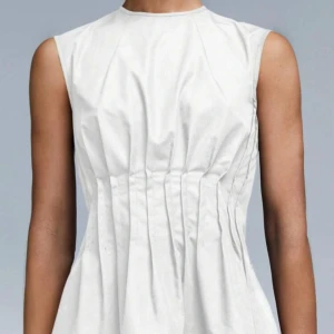 Vit ärmlös blus med knappar bak - Snygg vit blus i bomull med ärmlös design och markerad midja. Baksidan har en djup slits och svarta knappar som ger en cool kontrast. Perfekt till både jeans och kjol för en clean look.