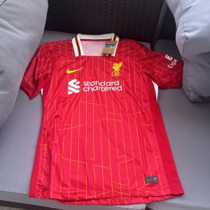 Liverpool FC röd matchtröja Nike M - Liverpool FCs officiella matchtröja från Nike i rött med gula ränder och klubbmärke på bröstet. Tröjan har vit krage med röda och gula detaljer, korta ärmar och är tillverkad i lätt, ventilerande polyester. Snygg design med sponsortryck och klassiska klubbfärger.
