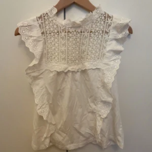 Vit blus med spets och volang - Supersöt vit blus med broderad spets upptill och volangdetaljer längs axlar och fram och bakstycke. Ärmlös modell  och rund halsringning. 