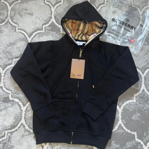 Svart Burberry hoodie med dragkedja - Svart hoodie från Burberry i bomull med klassiskt rutigt foder i huvan. Tröjan har hel dragkedja i guld, snörning i huvan och två fickor framtill. Perfekt för dig som vill ha en stilren och lyxig streetwear-look. Priset kan diskuteras, köparen står för frakten.