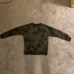 Grön camo crewneck sweatshirt - Säljer en crewneck sweatshirt i mörkgrön camouflage med svarta detaljer. Tröjan har rund halsringning, långa ärmar och muddar vid ärmslut och nederkant. Perfekt för dig som gillar streetwear och vill sticka ut lite extra.