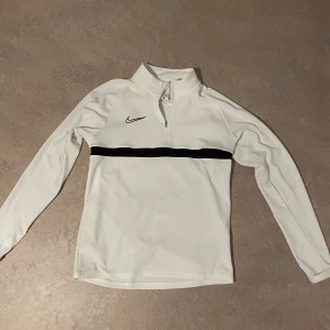 Vit Nike half zip tröja med svart detalj - Snygg vit långärmad tröja från Nike med svart horisontell rand över bröstet och klassisk Nike-logga. Tröjan har half zip och hög krage. Tillverkad i mjukt och stretchigt syntetmaterial. I nytt skick. Används ett oerhört fåtal gånger.
