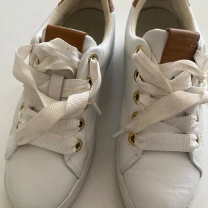 Vita sneakers från Gant med bruna detaljer - Snygga vita sneakers från Gant med breda vita snören, guldfärgade öljetter och bruna skinnpaneler på häl och plös. Skorna är i skinn och har en klassisk, rund tå och platt sula. Perfekta för en clean och stilren look. Använda ett fåtal ggr, mycket bra skick! 