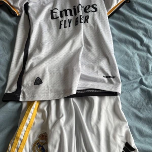 Real Madrid fotbollströja Adidas vit - Real Madrid fotbollströja från Adidas i vit med svarta och gula detaljer. Tröjan har korta ärmar, svart Adidas-logga och klubbmärke på bröstet samt 'Emirates Fly Better' tryckt framtill. Tillverkad i lätt och ventilerande polyester med AEROREADY-teknologi. Det finns vini.jr och benzema pris kan oxå diskuteras för ett rimligare pris. 