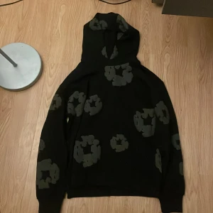 Svart hoodie från Denim Tears med mönster - Svart hoodie från Denim Tears med unika gråa mönster över hela tröjan. Oversized fit med huva och långa ärmar. Tillverkad i USA och perfekt för dig som gillar streetwear med edge.