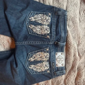 Miss Me jeans  - Mörkblå jeans från Miss Me med snygga broderade detaljer i vitt och silver på bakfickorna. Klassisk bootcut-modell med låg midja och slitna kanter nertill. innerbenslängd:81cm midjemått:38cm