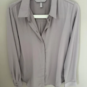 Ljusgrå skjorta från H&M - Stilren ljusgrå skjorta från H&M med klassisk krage och knappar framtill. Skjortan har lång ärm med manschett och en rak passform, perfekt för en clean och enkel look. Tillverkad i ett mjukt och lätt material som känns skönt mot huden.