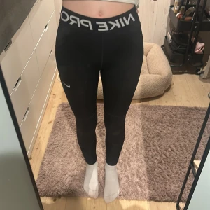 Svarta Nike Pro leggings  - Svarta Nike Pro leggings med hög midja och bred resår med logga. Tight passform och stretchigt material som passar perfekt till träning. Lite slitning bak vid midjan(se sista bilden) men inget hål, och Nike loggan är lite ur tvättad men inget som syns om man inte kollar nära!