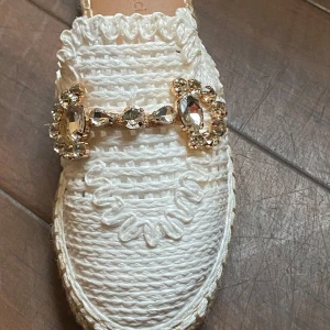 Vita espadrillos med strassdetalj - Snygga vita espadrillos i stickat material med dekorativ strassdetalj i guld framtill. Skon har en platt sula i klassisk jute och en rund tå. Perfekt för dig som vill ha en unik och trendig sommarsko med extra bling.