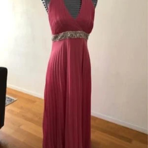 Rosa långklänning med paljettbälte - Elegant rosa långklänning med v-ringad omlott-topp och markerad midja med glittrigt paljettbälte. Klänningen har plisserad kjol och är ärmlös, perfekt för bal eller festligare tillfällen.