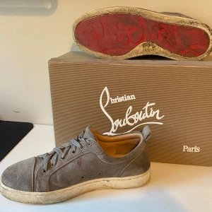 Grå mockasneakers från Christian Louboutin - Sparsamt använda med en lite röd fläck som syns.    Snygga grå sneakers från Christian Louboutin i mocka med klassisk röd sula. Skorna har rund tå, snörning och platt sula. Insidan är fodrad i beige läder och utsidan har en diskret röd detalj. Perfekta för dig som gillar exklusiva och stilrena sneakers.