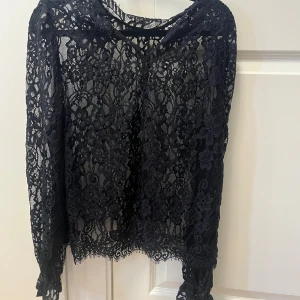 Svart spetsblus med lång ärm - Säljer en svart blus i genombruten spets med blommigt mönster. Blusen har långa ärmar och rund halsringning. Perfekt för dig som vill ha en elegant och trendig look med mycket detaljer. Spetsen ger en lyxig känsla och blusen är lätt att styla till olika outfits.