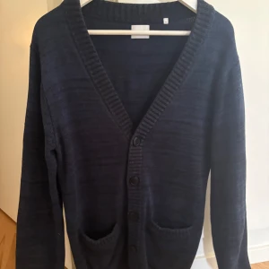 Stickad cardigan  - Mysig mörkblå kofta från Bläck i storlek M. Koftan har djup v-ringning, knäppning med fyra knappar och två fickor framtill. Stickad i mjukt material. 