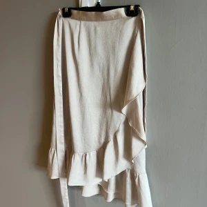 Beige omlottkjol med volang Neo Noir - Supersöt beige omlottkjol från Neo Noir med volangkant och knytband i midjan. Kjolen har en lätt och flowy känsla, perfekt för varmare dagar. Stilren och enkel att matcha med både sneakers och klackar. 