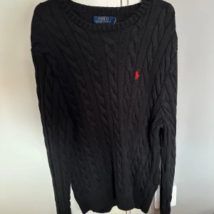 Svart kabelstickad tröja Polo Ralph Lauren - Kabelstickad svart tröja från Polo Ralph Lauren med klassisk röd logga broderad på bröstet. Tröjan har rund halsringning och långa ärmar, perfekt för lager-på-lager-stil under kyliga dagar.