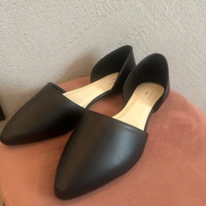 Svarta ballerinaskor från Nelly - Helt nya endast testade svarta ballerinaskor från Nelly med spetsig tå och platt sula. Skorna har en minimalistisk design i syntetmaterial och öppen insida, vilket ger en modern och clean look. Perfekta för dig som gillar enkel och trendig stil.