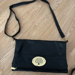 Svart axelväska från Mulberry - Snygg svart axelväska från Mulberry i skinn med justerbar rem och guldfärgade metalldetaljer. Väskan har ett stort fack med dragkedja och en dekorativ Mulberry-logga framtill. Perfekt för dig som gillar stilrena accessoarer med lyxig känsla.