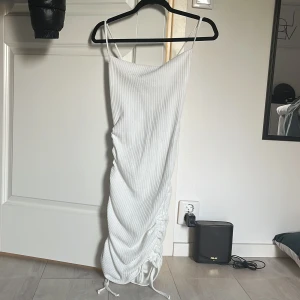 Vit ribbad långklänning med snörning - Supersnygg vit ribbad långklänning från prettylitlething med smala axelband och trendig snörning längs ena sidan. Klänningen har en tight passform och är tillverkad i mjuk bomullsmix. Perfekt för dig som vill sticka ut med en stilren look.