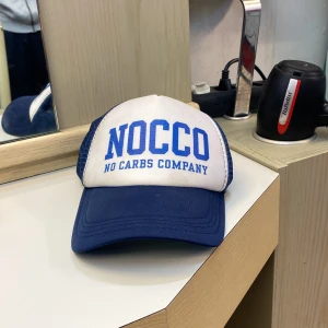 Blå och vit NOCCO keps - Snygg truckerkeps från NOCCO med vit frontpanel och blått tryck, samt blått nät på sidor och bak. Böjd skärm i blått och justerbar snapback. Perfekt för dig som gillar sportig och avslappnad stil. Skick: 9/10