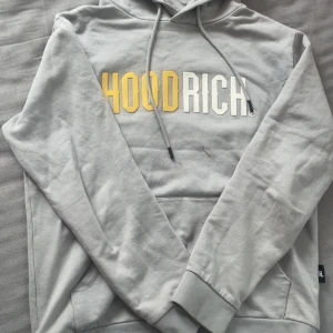 Grå Hoodrich hoodie med tryck - Snygg grå hoodie från Hoodrich med stor HOODRICH-logga i gult och vitt framtill. Tröjan har huva med snörning och en klassisk känguruficka. Perfekt för dig som gillar streetwear och vill ha en stilren look.