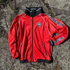 Röd track jacket från Umbro - Röd track jacket från Umbro med svartvita detaljer längs ärmarna och broderad logga på bröstet och ryggen. Jackan har hel dragkedja, höga muddar och två fickor med dragkedja. Perfekt för dig med sportig och retro stil!                   Mått: Chest (A): 47cm (B): 63cm , Arms: 56cm