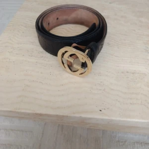 Svart Gucci skärp med GG-spänne - Snyggt svart skärp från Gucci i äkta skinn med klassiskt guldigt GG-spänne. Bältet har diskret präglat mönster längs hela remmen och insidan är i naturfärgat läder. Perfekt accessoar för att lyfta din outfit med en lyxig touch.