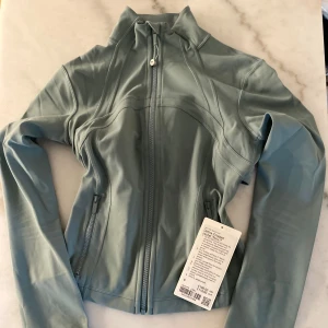 Define Cropped Jacket från Lululemon - Snygg ljusblå Define Cropped Jacket från Lululemon i mjukt och stretchigt Nulu-material. Jackan har slim fit, dragkedja framtill, zip-fickor och tumhål i ärmsluten. Perfekt för träning eller chill, med en clean och sportig look.