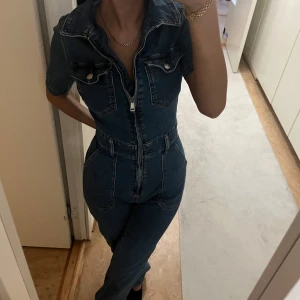 Jeans jumpsuit med dragkedja - En snygg jeans jumpsuit i blå denim med korta ärmar, markerad midja och dragkedja framtill. 