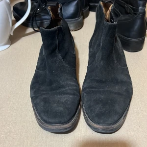 Svarta mocka chelsea boots från Gant - Svarta chelsea boots från Gant i mjuk mocka med elastiska paneler på sidorna och dragflik bak. Klassisk rund tå och platt sula med diskret Gant-logga på sidan. Perfekta för en stilren och enkel look.