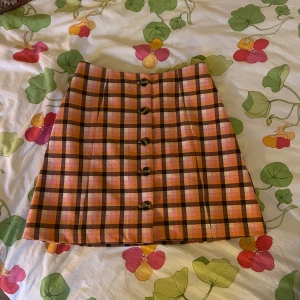 Rutig kortkjol med knappar - Superfin kortkjol i rutigt mönster med rosa, orange och bruna toner. Kjolen har knappar framtill som snygg detalj och en klassisk A-linjeform. Perfekt för dig som vill sticka ut med färg och mönster.