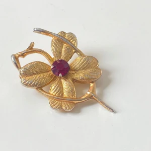 Vintage brosch från Coro i guldton - Vintage brosch från Coro i form av en blomma med fyra blad i guldtonad metall och en röd sten i mitten. Bladens yta har fina detaljer och broschen har en klassisk nål på baksidan. Perfekt accessoar för att ge din outfit en unik vintagekänsla.