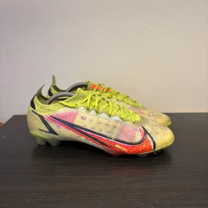 Nike Mercurial fotbollsskor gul/röd storlek 44 - Nike Mercurial fotbollsskor i neon-gult med röda och svarta detaljer. Skorna har snörning, låg profil och är tillverkade i syntetmaterial för lätt känsla på planen. Swoosh-logga på sidan och mönstrad ovandel för extra grepp. Perfekta för dig som vill sticka ut på fotbollsplanen.