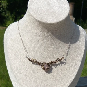 Handgjort halsband med bronsfärgad tråd - Unikt halsband med tunn silverkedja och ett detaljerat hänge i bronsfärgad tråd, formad i snirkliga mönster runt en ljusrosa hjärtformad sten. Perfekt statement-piece för dig som gillar boho och romantiska vibes.