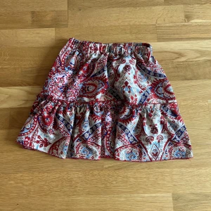 Röd och blå mönstrad kortkjol XS - Kortkjol i storlek XS med färgstarkt paisleymönster i rött, blått och vitt. Kjolen har volangdetalj och resår i midjan för en bekväm passform. Perfekt för dig som gillar boho-vibe och vill sticka ut med coola mönster.