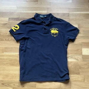 Ralph lauren piké  - Klassisk marinblå pikétröja från Polo Ralph Lauren med gul broderad logga och nummerdetalj på ärmen. Tröjan har krage, korta ärmar och knappar vid halsen. Tillverkad i bomull och passar perfekt för en sportig och stilren look.