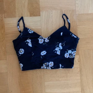 Blommig croppad linnetopp - Svart/mörkblå croppad linnetopp med smala axelband och prästkrageblommor. Toppen har v-ringning och knytning i ryggen för extra detalj. 