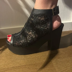 Svarta platåklackar med spets från Vox - Snygga svarta platåklackar från Vox med öppen tå och grov klack. Skorna har detaljer i spets och justerbart spänne vid ankeln. 