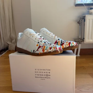 Maison Margiela sneakers med färgstänk - Unika vita sneakers från Maison Margiela med färgstänk i rött, orange, blått, svart och grönt. Skorna har klassisk låg siluett, snörning och brun sula. Tillverkade i skinn och perfekta för dig som vill sticka ut med en kreativ vibe.